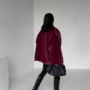 May include: A person wearing a burgundy coat, black leggings, and black knee-high boots. A black, gathered-style handbag is held in the hand. The background is a plain white wall and floor.