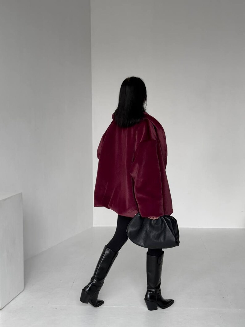 May include: A person wearing a burgundy coat, black leggings, and black knee-high boots. A black, gathered-style handbag is held in the hand. The background is a plain white wall and floor.