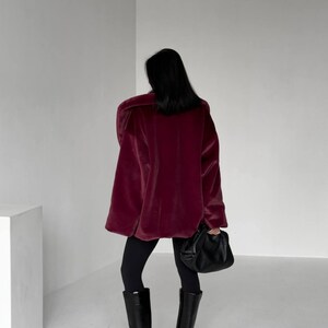 May include: A person wearing a burgundy oversized coat, black leggings, and black knee-high boots. The person is holding a black handbag. The coat has a plush, velvety texture. The boots have a pointed toe and a block heel.