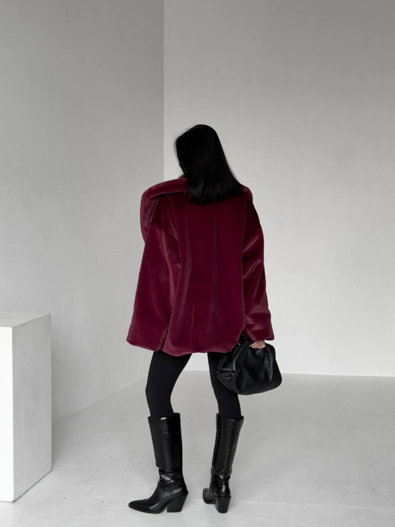 May include: A person wearing a burgundy oversized coat, black leggings, and black knee-high boots. The person is holding a black handbag. The coat has a plush, velvety texture. The boots have a pointed toe and a block heel.