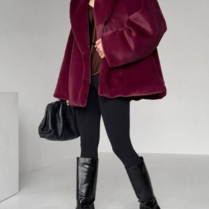 May include: A burgundy faux fur coat with a wide collar and long sleeves. The coat is paired with black leggings and knee-high black leather boots. A black handbag is held in the left hand. The outfit is set against a white background.