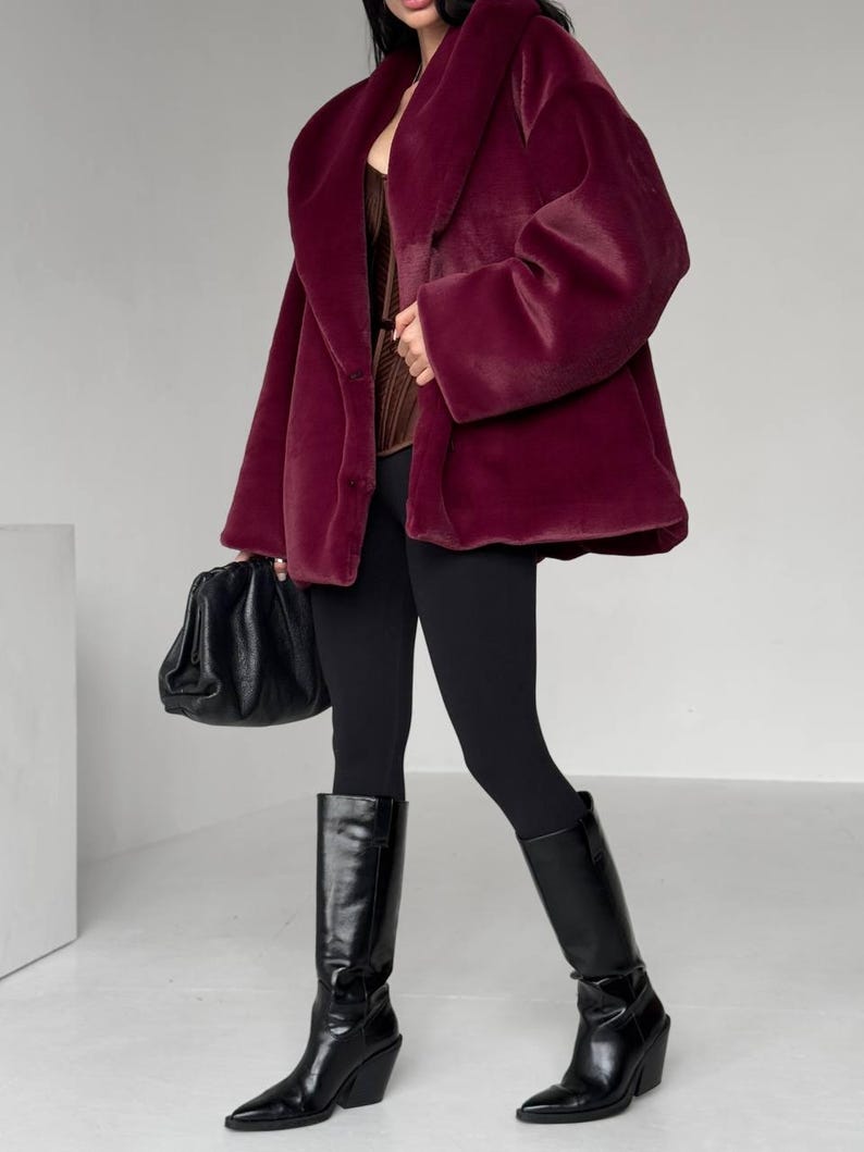 May include: A burgundy faux fur coat with a wide collar and long sleeves. The coat is paired with black leggings and knee-high black leather boots. A black handbag is held in the left hand. The outfit is set against a white background.