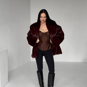 May include: A woman wearing a burgundy faux fur coat over a brown corset top, black leggings, and black knee-high boots. The coat has a wide collar and the boots have buckles. The outfit is set against a plain white background.