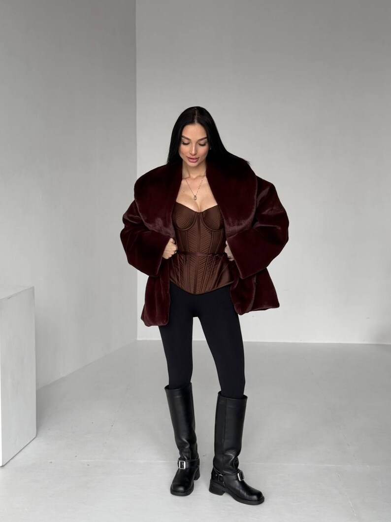 May include: A woman wearing a burgundy faux fur coat over a brown corset top, black leggings, and black knee-high boots. The coat has a wide collar and the boots have buckles. The outfit is set against a plain white background.