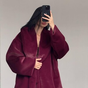 May include: A burgundy faux fur coat with a wide collar and long sleeves. The coat is buttoned and appears soft and plush. The coat is worn over a dark top and black pants.