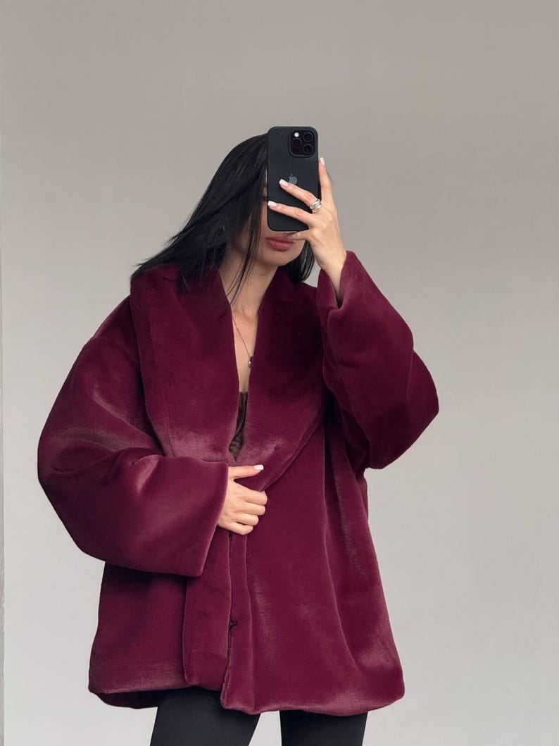 May include: A burgundy faux fur coat with a wide collar and long sleeves. The coat is buttoned and appears soft and plush. The coat is worn over a dark top and black pants.