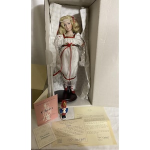 May include: A porcelain doll named "Clara" from The Nutcracker Doll Collection, wearing a white dress with red accents and matching shoes. The doll has blonde hair and is displayed in its original box with accompanying paperwork.