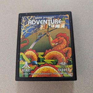 May include: A black rectangular Atari video game cartridge for the game Adventure. The label features a colorful illustration of a red dragon, a castle, and a path. Text on the label includes game program, ADVENTURE, ATARI, and CX2613 1978.
