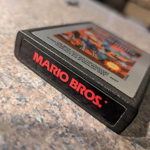 May include: A black Nintendo game cartridge for Mario Bros. The game cartridge has a red label with the words "MARIO BROS." and a colorful image of the game on the top. The cartridge is resting on a speckled surface.
