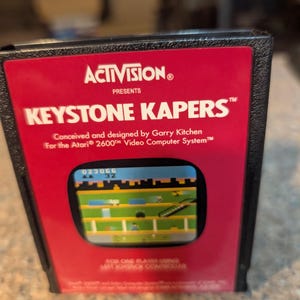 May include: A black plastic Atari 2600 video game cartridge for Keystone Kapers by Activision. The red label features white text reading Activision Presents Keystone Kapers, Conceived and designed by Garry Kitchen for the Atari 2600 Video Computer System, and a small screen capture of the game.