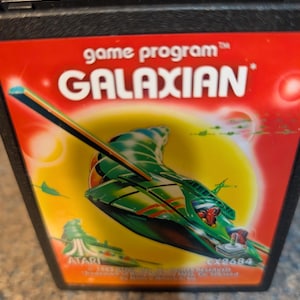 May include: A vintage Atari Galaxian game cartridge. The label features a green and red spaceship design with the game title in white text. The background is yellow and red with the Atari logo.