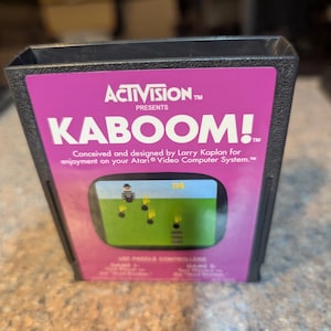 May include: A black plastic Atari video game cartridge for the game Kaboom! by Activision. The label is magenta with white text reading Activision Presents Kaboom! and includes a small screen capture of the game showing a character and bombs.