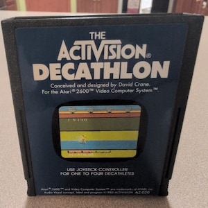 May include: A black Atari 2600 video game cartridge for The Activision Decathlon. The label features white text, the title, and a small screen capture of the game showing a runner on a track. It includes instructions to use a joystick controller.