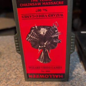 May include: A vintage video game cartridge for "The Texas Chainsaw Massacre" and "Halloween" from Wizard Video Games. The cartridge has a red label with black text and a graphic. The game is labeled No. 007 and No. 008.
