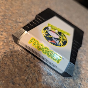 May include: A black plastic video game cartridge for the Atari 2600 console. The white label features the green text FROGGER and a circular graphic of a frog. Small black text at the bottom includes copyright information for Sega Enterprises.