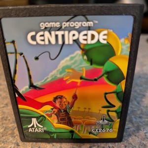May include: A vintage Atari Centipede game cartridge. The label features the game title in large white letters, with a green centipede and a character holding a weapon. The Atari logo is visible, along with the code CX2676.
