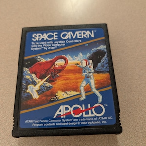 May include: A black rectangular video game cartridge for the Atari 2600 titled Space Cavern by Apollo. The label features a blue and orange illustration of an astronaut fighting a red creature on a planet surface with text including 1982 copyright.