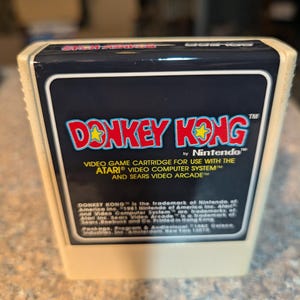 May include: A beige plastic video game cartridge for the Atari Video Computer System. The label features the red Donkey Kong logo with yellow stars, white text stating it is for use with Atari and Sears systems, and legal copyright information.