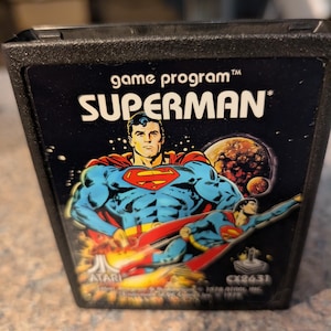 May include: Black Atari game cartridge for the game "Superman." The label features a comic book style illustration of Superman in his iconic blue and red suit, with the words "game program" and "SUPERMAN" in white text.