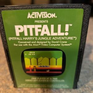 May include: A black Atari video game cartridge for Pitfall! featuring a green label with white text that reads Activision Presents Pitfall! Pitfall Harry's Jungle Adventure, designed by David Crane for the Atari Video Computer System.