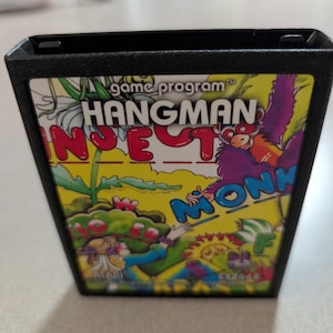May include: A black plastic Atari video game cartridge for the game Hangman. The label features a colorful illustration with yellow, red, and blue accents, displaying the text game program HANGMAN, along with partial words and graphics.
