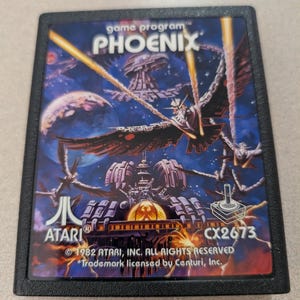 May include: A black Atari game cartridge for the game Phoenix. The label features a blue space scene with a large bird-like spaceship, planets, and yellow laser beams. Text includes game program, PHOENIX, ATARI, CX2673, and 1982 Atari, Inc.