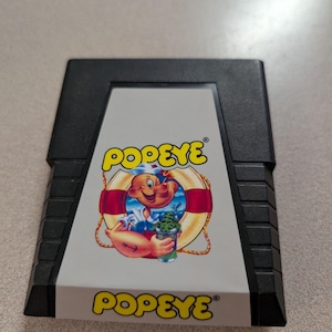 May include: A black video game cartridge for the Nintendo Entertainment System featuring a label with the word POPEYE in yellow letters. The label displays a cartoon illustration of Popeye the Sailor holding a can of spinach inside a life preserver.