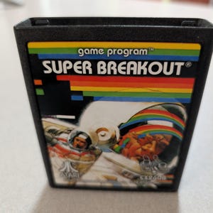 May include: A black Atari video game cartridge for Super Breakout. The label features a colorful rainbow graphic, the text game program, Super Breakout, and Atari, along with an illustration of a person in a space suit and a bowl of colorful blocks.