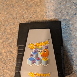May include: A black video game cartridge for the Atari 2600 featuring a gray label with the Q*bert logo in yellow and orange. The label displays the orange Q*bert character on a blue isometric pyramid structure with purple enemies and a red ball.