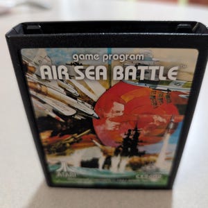 May include: A black Atari video game cartridge for the Air-Sea Battle game program. The label features a colorful illustration of military aircraft and naval ships in combat, with the title text Air-Sea Battle printed in white at the top.