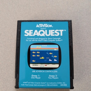 May include: A black and blue Atari video game cartridge for Seaquest by Activision. The label features the game title, designer credit, and a screenshot of the underwater gameplay. Text includes 'Activision', 'Seaquest', and 'Use Joystick Controllers'.