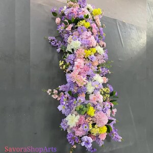 May include: A long, vertical floral arrangement with pastel-coloured flowers. The arrangement includes pink, purple, yellow, and white blooms, with green foliage. The text "SavoraShopArts" is visible at the bottom.