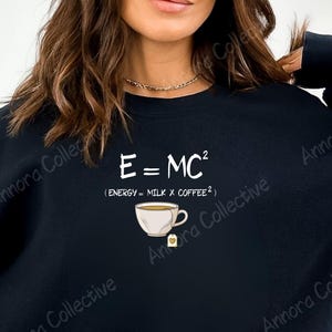 May include: Black sweatshirt with the text "E=MC²" and the equation "(ENERGY = MILK X COFFEE²)". A white teacup graphic is below the text. The sweatshirt is a casual style.
