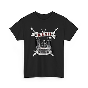 May include: Black t-shirt featuring a white and red crest design. The crest includes a ship, towers, and the word "KESH" in a banner. The text "EST. CORCAIGH 2014" is below the crest. Two crossed guitars are on the sides.