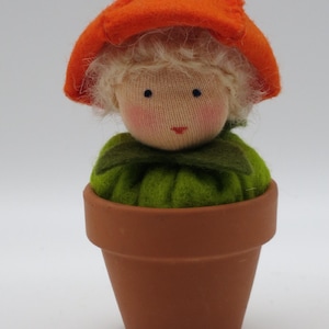 Op de afbeelding: Een vilten pop met blond haar, die een oranje pompoenhoed en een groene jurk draagt, zit in een terracotta bloempot.