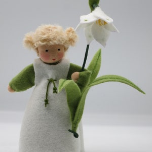 Könnte beinhalten: Handgemachte Filzpuppe mit blonden Locken, die ein weißes Kleid und grüne Ärmel trägt. Die Puppe hält eine große weiße Blume mit grünen Blättern und einem schwarzen Stiel. Die Puppe hat ein bemaltes Gesicht mit rosigen Wangen.