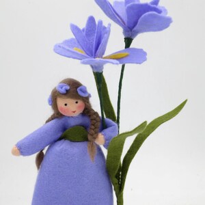 May include: A handcrafted felt doll with brown braided hair, wearing a light purple dress and matching hair accessories, standing next to two tall, vibrant purple felt flowers with green stems and leaves against a plain white background.