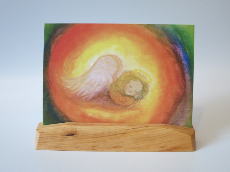 Op de afbeelding: Een houten standaard met een kleurrijke kaart met een schilderij van een engel met vleugels en een halo, die een slapend kind in een cirkel van licht vasthoudt.