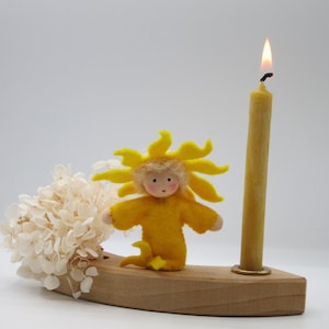 Pode incluir: Uma boneca de feltro amarela com cabeça em forma de sol e uma vela acesa em uma base de madeira. A boneca está sentada em uma base de madeira com uma flor de hortênsia branca atrás.