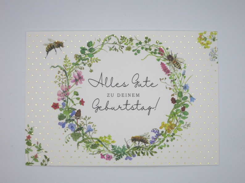 May include: A white card with gold polka dots and a wreath of flowers and bees. The text on the card reads "Alles Gute zu deinem Geburtstag!"
