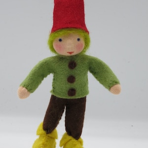 May include: A felt doll with a red hat, green shirt, brown trousers, and yellow shoes. The doll has blonde hair and a painted face.