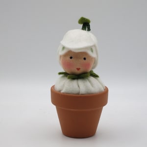 May include: A white felt doll with a green felt leaf on top of its head, wearing a white felt dress and sitting in a terracotta pot.