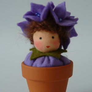 May include: A purple felt flower doll with brown hair sits in a terracotta flower pot. The doll has a green felt collar and blue beads on its flower crown.