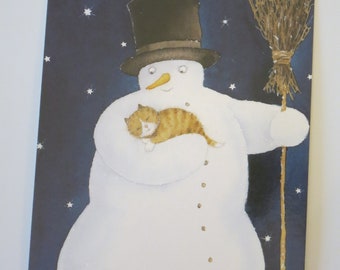 以下が含まれることがあります： 黒いシルクハットをかぶった雪だるまが、眠っているオレンジ色のトラ猫を抱いているグリーティングカード。雪だるまは人参の鼻とほうきを持っています。背景は白い星が散りばめられた濃い青色です。