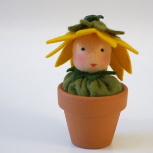 Könnte beinhalten: Eine Filzpuppe mit einem gelben Blumenkopf und grünen Blättern, die ein grünes Kleid trägt und in einem Terrakotta-Topf sitzt.