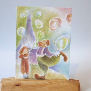 Op de afbeelding: Een aquarel schilderij van een clown die zeepbellen blaast terwijl een kind toekijkt. De clown draagt een paars en groen kostuum en een rode pruik. Het kind draagt een rode jurk en heeft rood haar. De achtergrond is een lichtblauwe en groene aquarel was met veel bellen.