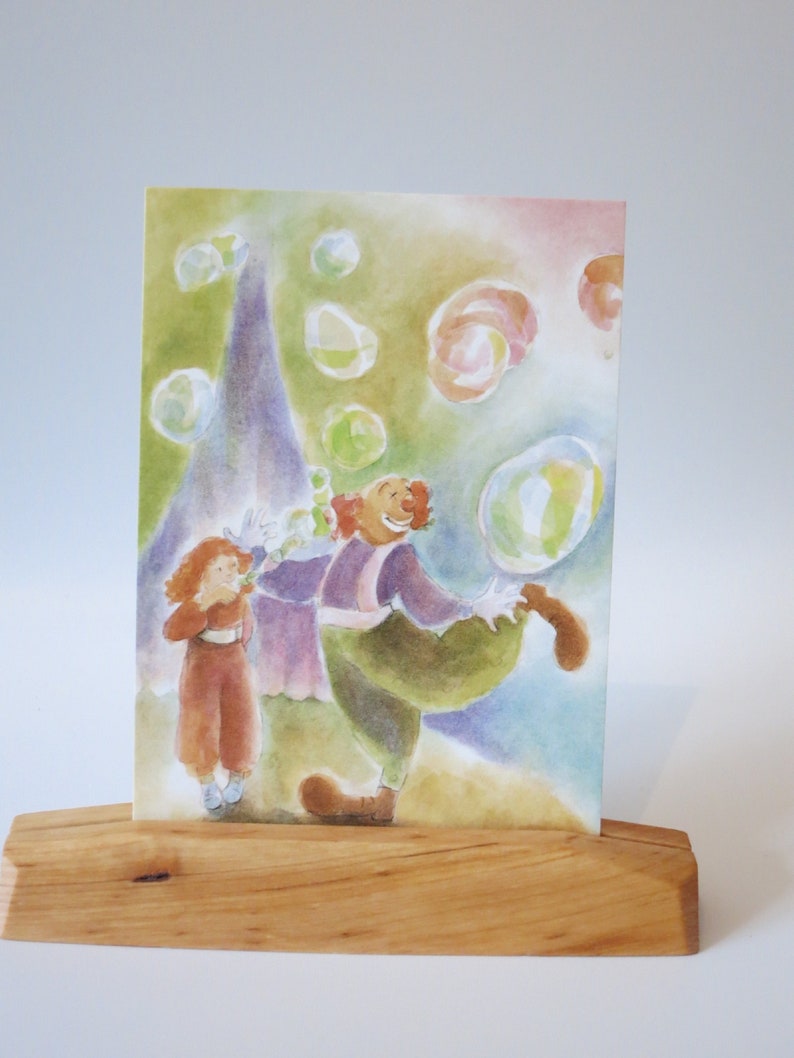 Op de afbeelding: Een aquarel schilderij van een clown die zeepbellen blaast terwijl een kind toekijkt. De clown draagt een paars en groen kostuum en een rode pruik. Het kind draagt een rode jurk en heeft rood haar. De achtergrond is een lichtblauwe en groene aquarel was met veel bellen.