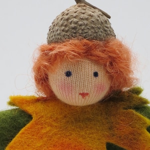 Op de afbeelding: Een handgemaakte vilten pop met rood haar die een eikelhoed en een groen en oranje bladkostuum draagt.