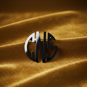 May include: A black metal brooch with the letters GNS in a circular design. The brooch is set against a gold velvet background, creating a contrast of textures and colors. The pin is a stylish accessory.