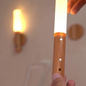 Puede incluir: Una luz LED cilíndrica de madera y blanca es sostenida en la mano de una persona. La luz tiene tres pequeños agujeros y un brillo cálido. El texto "HaveniqueDecor" está en la parte inferior de la imagen. Otra luz está montada en la pared.